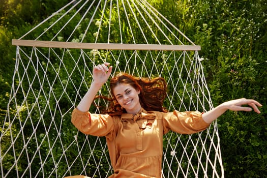 a beautiful, elegant woman lies in a long orange dress on a mesh hammock resting in nature, illuminated by the warm sunset light, smiling happily. High quality photo