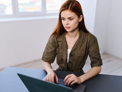 a woman sits at a table in a bright office with a laptop against the background of a window. High quality photo