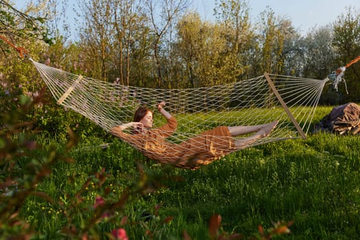 a happy woman in a long orange dress is relaxing in nature lying in a mesh hammock enjoying summer and vacation, looking at the sky. High quality photo