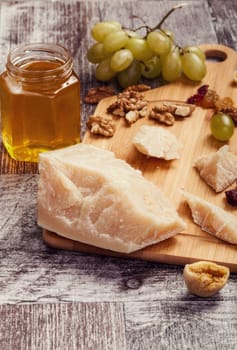 Big piece of parmesan next to nuts, grape and honey on vintage wooden background