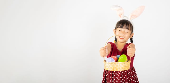 Happy Easter Day. Smile Asian little girl wearing easter bunny ears holding basket of full colorful eggs smiles broadly isolated on white background with copy space, Happy child in holiday