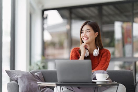 Beautiful Asian Woman smile and relaxing at home and using laptop computer sitting on cozy sofa.