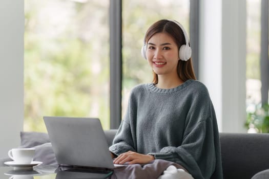 Beautiful Asian Woman smile and relaxing at home and using laptop computer sitting on cozy sofa.