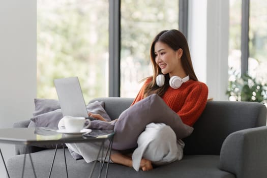 Beautiful Asian Woman smile and relaxing at home and using laptop computer sitting on cozy sofa.