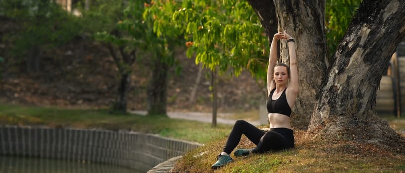 Happy athletic woman sitting in green park, warming up her arms before doing exercise. Fitness, training and healthy lifestyle concept.