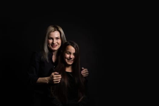 Young mother and her little daughter are looking at camera and smiling, sitting against black wall.