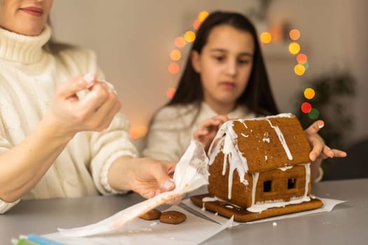 mother and daughter decorating gingerbread house. Beautiful living room with lights. Happy family celebrating holiday together