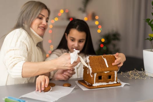 mother and daughter decorating gingerbread house. Beautiful living room with lights. Happy family celebrating holiday together