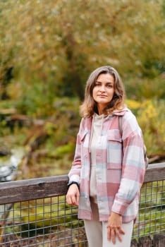 Portrait of cute young woman in casual wear in autumn, standing on bridge against background of an autumn Park and river. Pretty female walking in Park in golden fall. Copy space. smiling girl in the park standing on wooden bridge and looking at the camera in autumn season