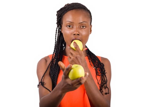 young girl standing in orange tank top on white background eating apple looking at camera.