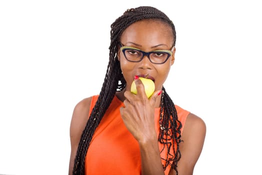 young girl standing in orange tank top on white background eating apple looking at camera.
