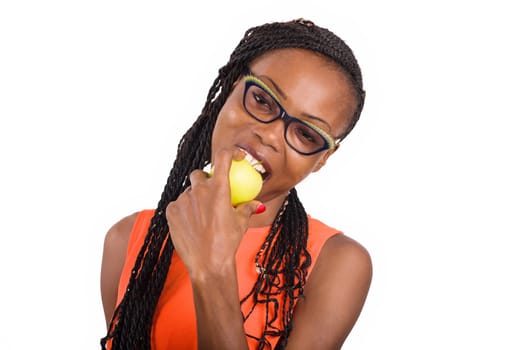 young girl standing in orange tank top on white background eating apple and looking at camera smiling.