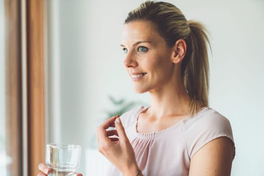 Smiling young caucasian woman taking her morning supplements, vitamin pills with a glass of water at home. High quality photo