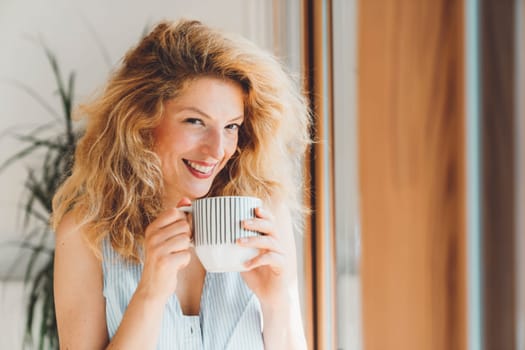 Cheerful happy caucasian woman relaxing at home on her day off work. Adult content woman spending time at home, in her apartment, resting on the couch, having a cup of coffee, feeling good.