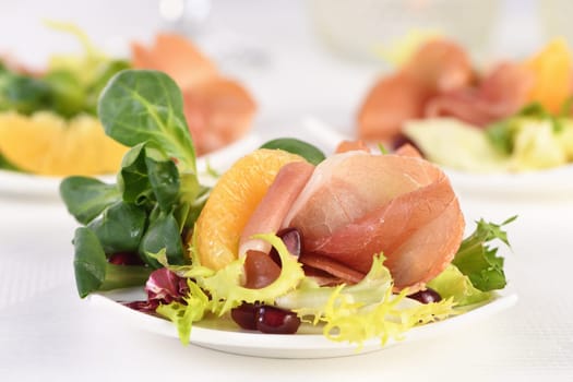 Portioned appetizer of prosciutto ham served on a of mixed with orange and pomegranate.