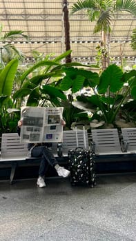 A Ukrainian refugee woman sits on a waiting bench and reads the newspaper New York Times with a suitcase at a train station with tropical palm trees. Spain Madrid