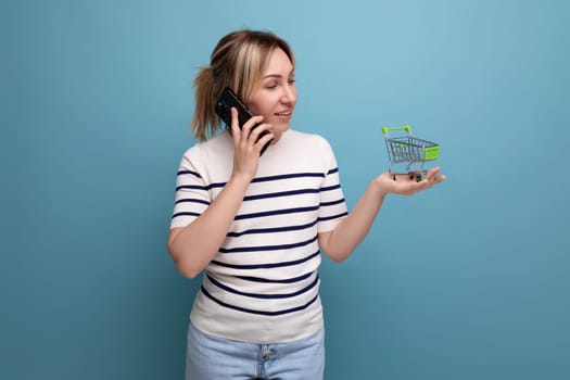 horizontal photo of attractive shopaholic girl talking on phone holding shopping cart on blue background with copy space.