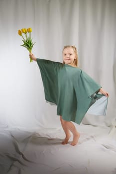 Portrait of a little blonde girl with bouquet of spring yellow flowers on a light background. Child in green dress holding a bouquet of tulips in hands. Spring concept