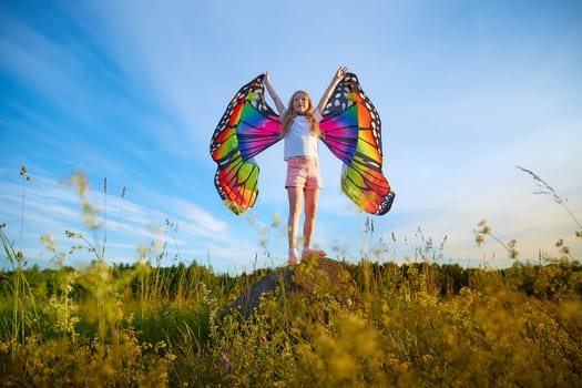 Pretty blonde girl with bright butterfly wings having fun in meadow on natural landscape with grass and flowers on sunny summer day. Portrait of teenage child in spring season outdoors on field