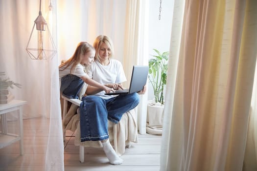 Beautiful young woman and her little cute daughter are using laptop at home. Enjoying spending time together with Internet and modern technologies