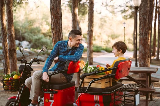 Farmer father and school boy kid delivered wooden crates with vegetables to camp site for picnic. Dad man male and son tourists delivered groceries on electric tricycle vehicle in the forrest