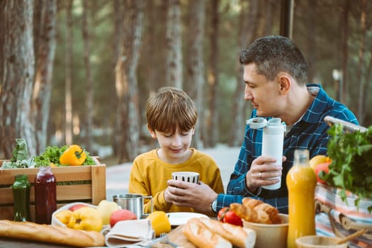 Father dad and school kid boy child having a picnic in the forest camping site with vegetables, juice, coffee, and croissants. Wooden crate with fresh organic veggies surrounded with bread baguettes