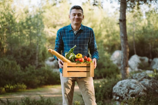 Caucasian man male holding wooden crate with vegan groceries organic vegetables and bread baguette. Farmer delivered a framed box with supplies to a farmers market camping site in the forest.