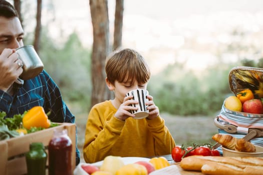 Father dad and school kid boy child having a picnic in the forest camping site with vegetables, juice, coffee, and croissants. Wooden crate with fresh organic veggies surrounded with bread baguettes