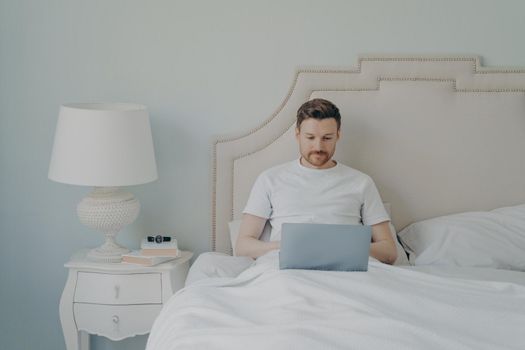 Attractive young european man watching video online on portable laptop computer while sitting in comfortable bed, using free internet connection at home. Leisure time indoor concept