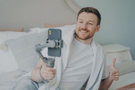 Young handsome caucasian bearded man doing online call with webcam using smartphone while lying on bed at home, happy with big smile doing super cool sign thumb up with fingers