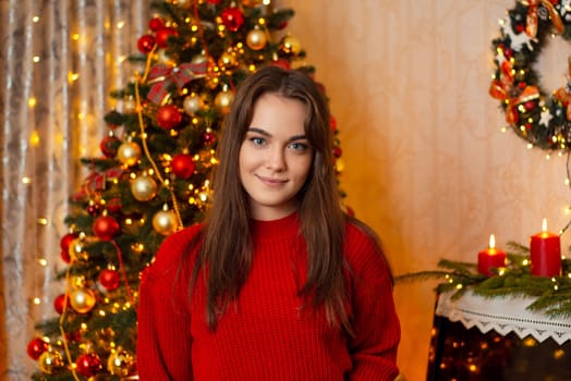 Portrait of a young attractive girl in red sweater looking to the camera, standing in decorated room with Christmas tree in the background