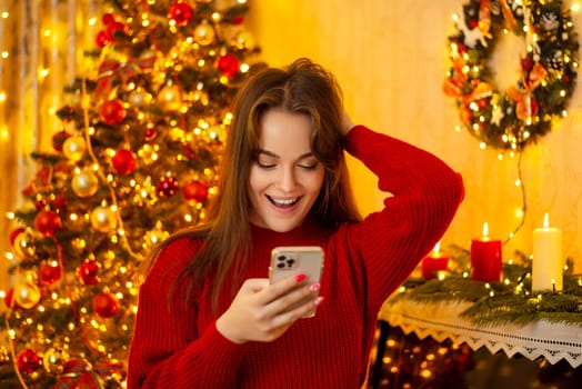 Amazed and surprised girl reading something on her phone, standing in decorated room with burning candles and a Christmas tree