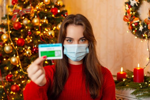 Girl in medical protective mask showing vaccination certificate to the camera standing in room with Christmas tree, candles, other decorations