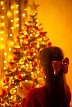 Atmospheric photo of a young girl admiring beauty of Christmas tree and decorations at home, drinking hot tea