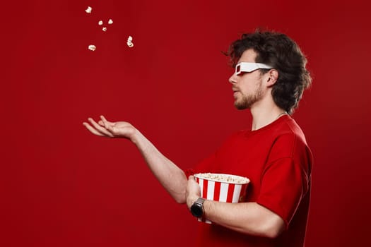 young bearded man in 3d-glasses, red t-shirt throwing up popcorn on red background, side view