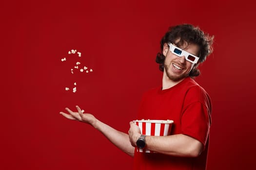 young happy man in 3d glasses, red t-shirt with flying popcorn on red background
