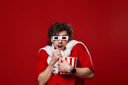scared handsome bearded man in 3d-glasses holding bucket of popcorn on red background