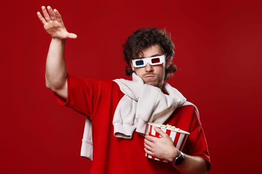 young bearded man in 3d-glasses holding big bucket of popcorn and dancing on red background