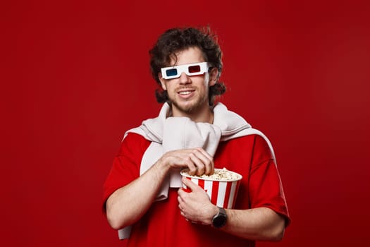 young bearded man in 3d-glasses eating popcorn on red background
