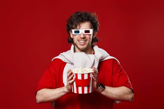 happy smiling bearded man in 3d-glasses holding big bucket of popcorns on red background