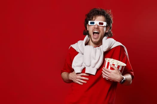 laughing bearded man in 3d-glasses, t-shirt with big bucket of popcorns on red background