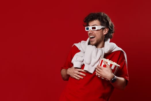 laughing bearded man in 3d-glasses, t-shirt with big bucket of popcorns on red background
