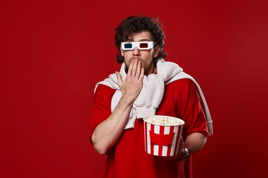 Shocked scared handsome man in 3d-glasses watching movie film hold bucket of popcorn on red background. copy space