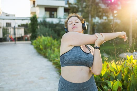 Adorable fat woman in tracksuit is engaged in fitness outdoor side view portrait. Young overweight woman lunges outdoors on warm summer day. Healthy lifestyle and weight loss.