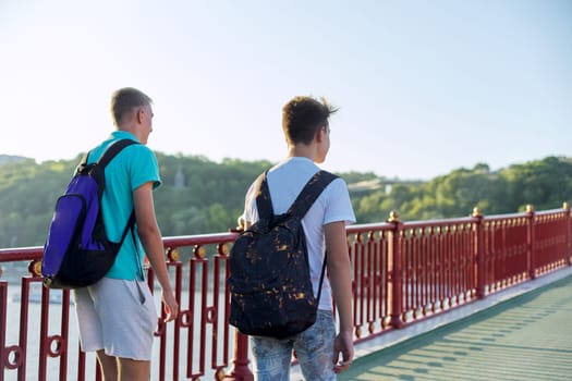 Two teenage boys with backpacks walking on bridge, back view, urban lifestyle, summer sunny day, with copy space