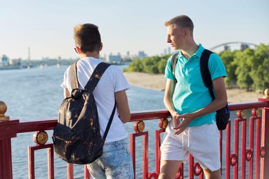 Outdoor portrait of two friends boys teenagers 15, 16 years old, talking laughing. Guys standing on bridge over river on sunny summer day. Youth, friendship, communication