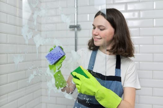 Bathroom cleaning, teenager girl washing shower glass with foam and sponges, closeup of females hand in green gloves and colored sponges, copy space