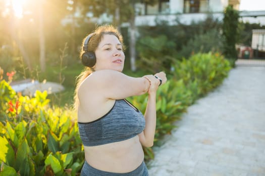 Adorable fat woman in tracksuit is engaged in fitness outdoor side view portrait. Young overweight woman lunges outdoors on warm summer day. Healthy lifestyle and weight loss.