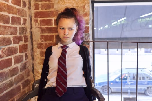 Portrait of 15 years old teenage girl in school uniform with tie sitting on chair, brick wall background, copy space window