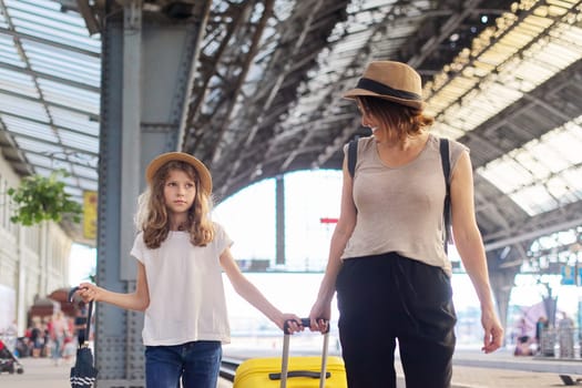 Happy mother and daughter child walking together at railway station with luggage suitcase. Travel, tourism, transportation, family concept